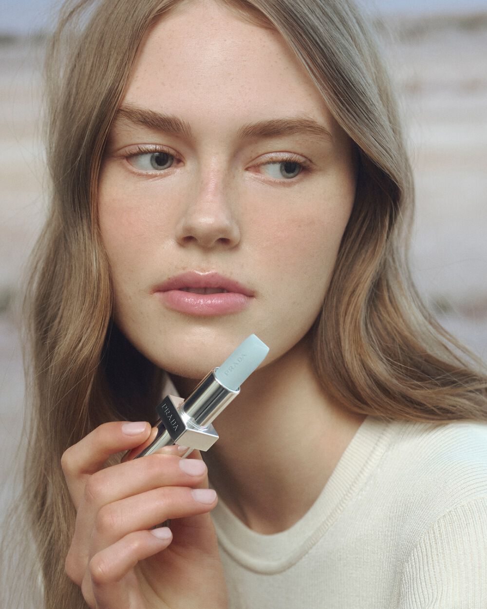 Prada Summer Stories Caucasian Model with Prada Balm in hand on a muted beach background 