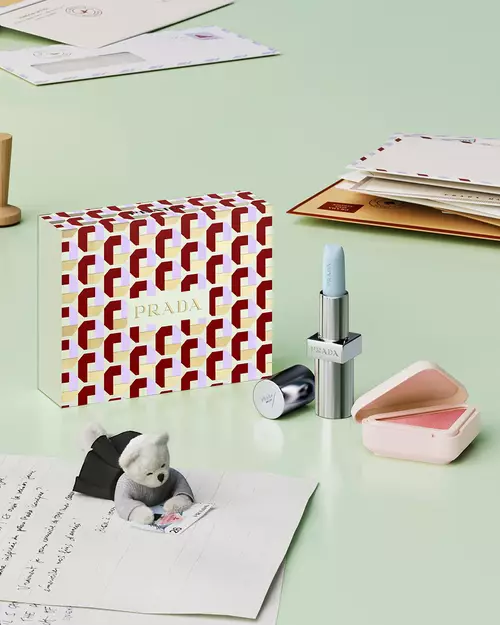 Prada Beauty Still Life visual with Holiday Beauty Clutch (mirror, mascara, lipstick, eye pencil and blush), letters, and teddy bear on a light green background