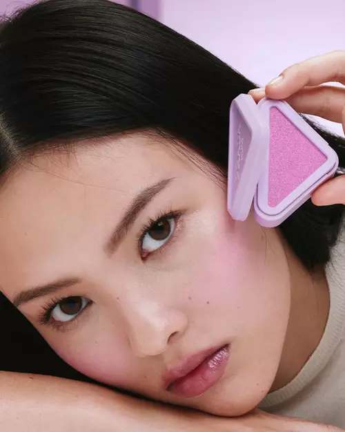 May Li (Asian skin model) is wearing Prada Touch Blush in shade P36 Wisteria, Pradascope Mascara in shade 01 Nero, and Prada Balm in shade U003 Wisteria Gleams on a lilac background.