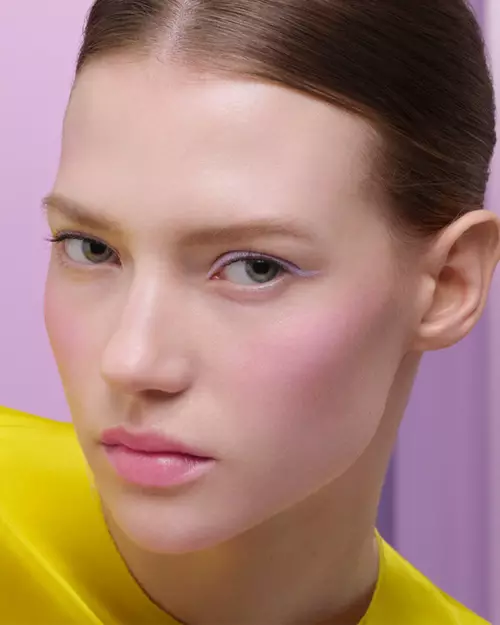 Olga (Caucasian Model) is wearing Prada Balm in shade Wisteria Gleams, Prada Lines Eye Pencil in shade Glycine, Pradascope Mascara in shade Nero, and Prada Touch Blush in shade Wisteria on a lilac background.