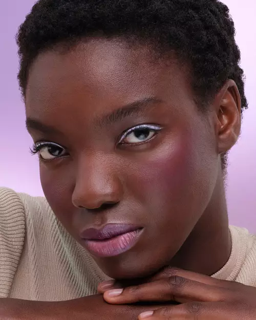 Ama (Deep skin model) is wearing Prada Touch Blush in shade P37 Violet, Prada Lines Eye Pencil in shade 06 Glycine, Pradascope Mascara in shade 01 Nero, and Prada Balm in shade U003 Wisteria Gleams on a lilac background.