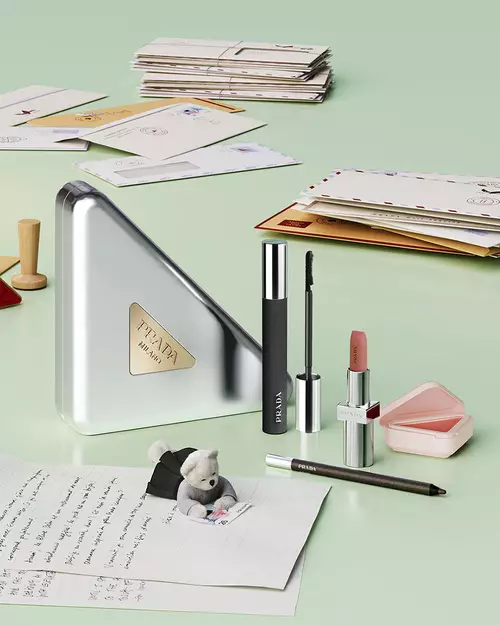 Prada Beauty Still Life visual with Holiday Beauty Clutch (mirror, mascara, lipstick, eye pencil and blush), letters, and teddy bear on a light green background