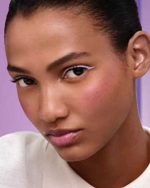 Cristi (medium skin model) is wearing Prada Balm in shade Wisteria Gleams, Prada Lines Eye Pencil in shade Glycine, Pradascope Mascara in shade Nero, and Prada Touch Blush in shade Violet on a lilac background.