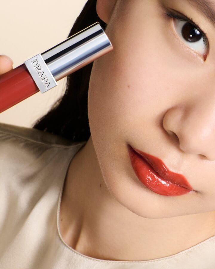 Model with East Asian features wearing the Prada Reflection Glossy Liquid Balm in shade Infrared while holding the liquid balm tube next to her face.