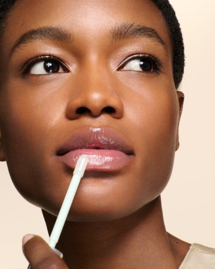 Model with deep skin tone applying the Prada Reflection Glossy Liquid Balm in the Universal shade.