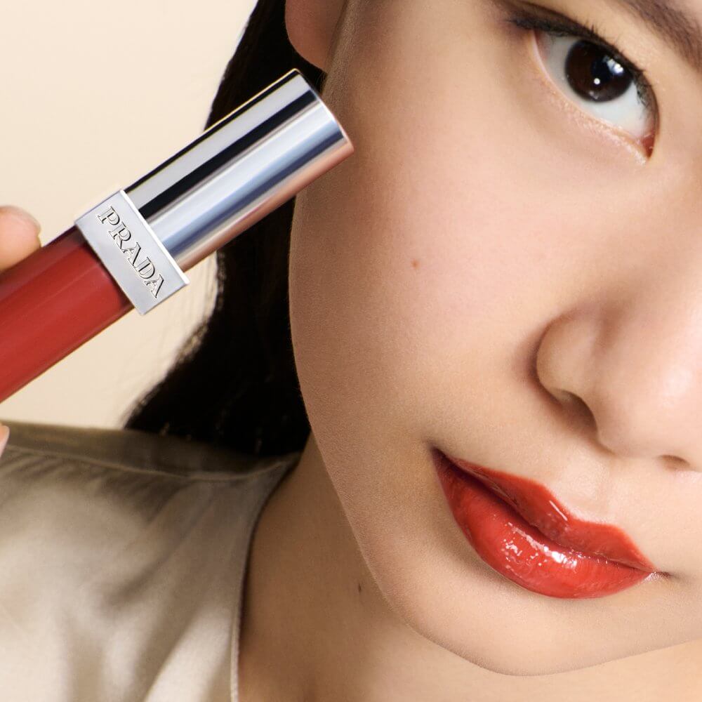 Model with East Asian features wearing the Prada Reflection Glossy Liquid Balm in shade Infrared while holding the liquid balm tube next to her face.